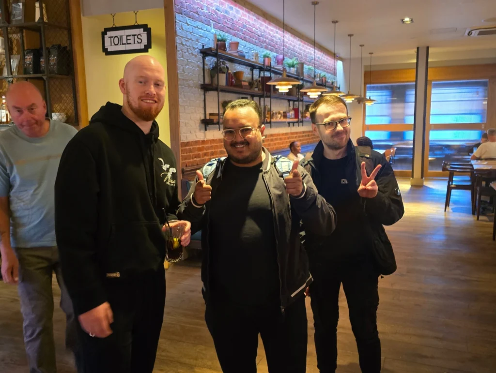 A-group-of-three-men-posing-in-a-restaurant,-smiling-and-giving-thumbs-up-with-another-man-in-the-background-walking-toward-the-restroom