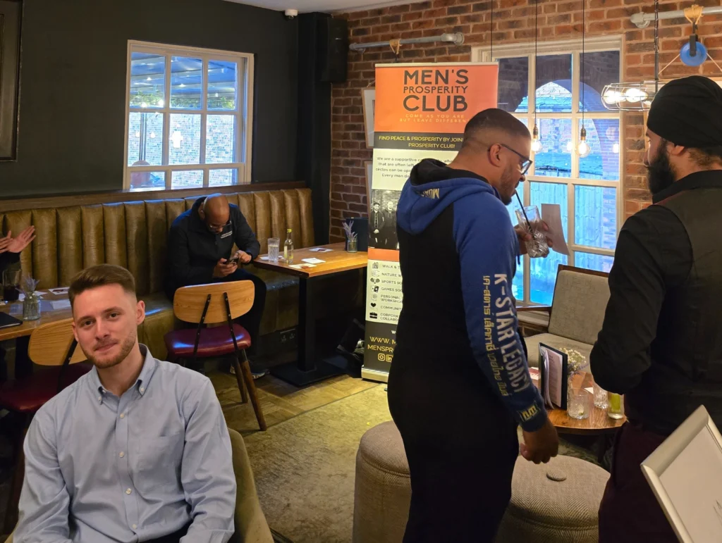 A-man-seated-at-a-table,-with-others-standing-in-the-background,-all-at-the-Men's-Prosperity-Club-event