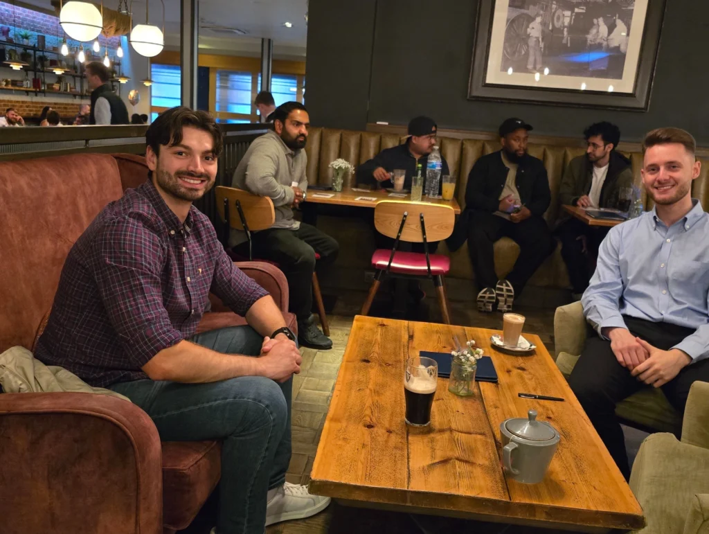 Two-men-sitting-at-a-table-in-a-cafe,-smiling,-with-other-members-in-the-background