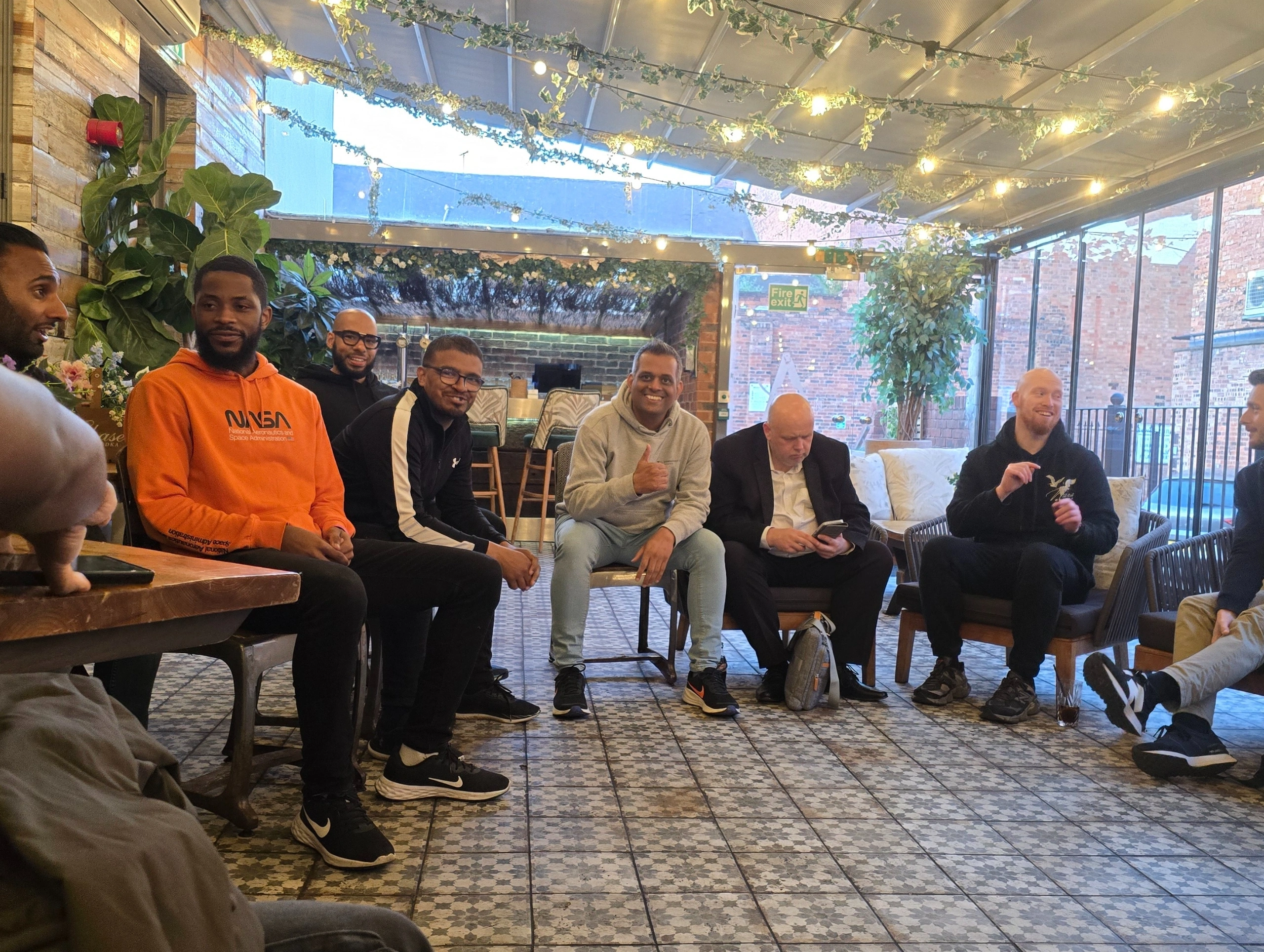 Two-men-sitting-at-a-table,-smiling-and-enjoying-a-conversation-in-a-warm,-cozy-café-environment-with-plants-around
