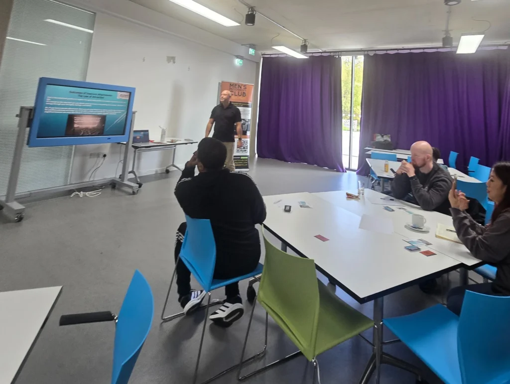 A-PD-workshop-by-John-Kenny-with-the-presenter-standing-and-discussing-attachment-theory-while-the-audience-listens-at-a-table